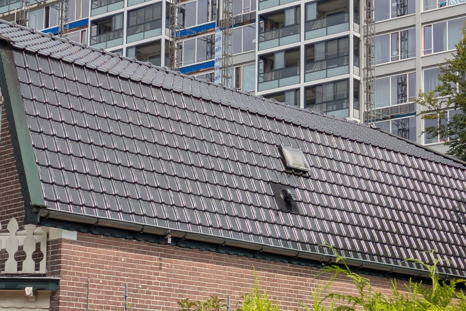 Een nieuw dak van donkere pannen met een dakraam; erachter een flat