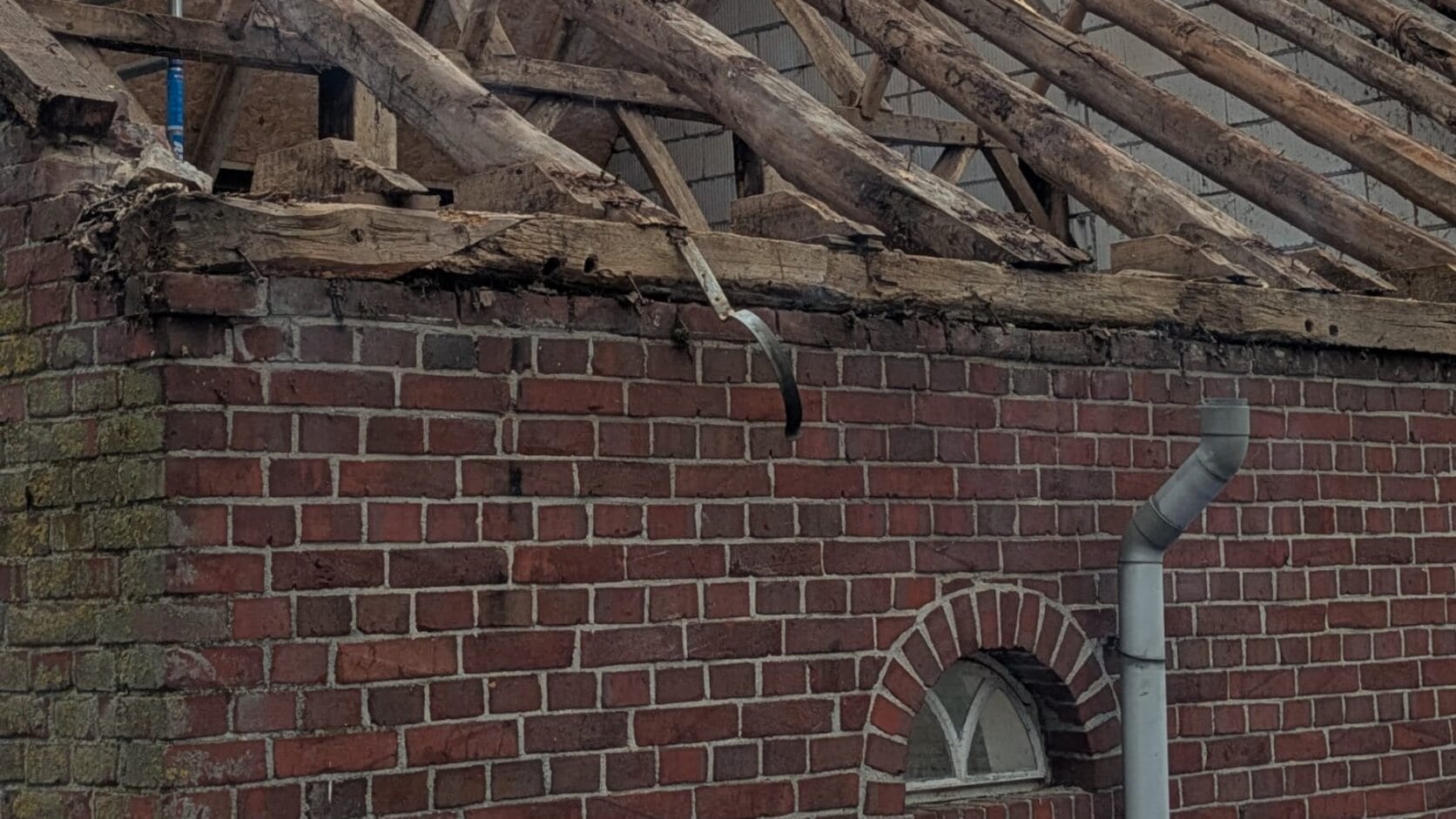 De bakstenen muur van een schuur met een rond raampje; het dak erboven is eraf